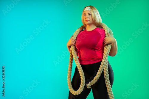 Young caucasian plus size female model's training on gradient green background in neon light. Doing workout exercises with the ropes. Concept of sport, healthy lifestyle, body positive, equality.