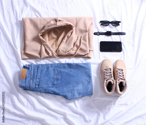 Blue jeans, beige sweatshirt,beige sneakers,black sunglasses, mobile phone and wrist watch on white sheet on bed. Overhead view of woman's casual day outfits. Trendy hipster look. Flat lay.