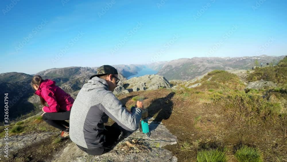 A young couple cooking in the wilderness, nearby of Preikestolen, Norway. Wild camping in the nature. The girl collects the wood, the man cooks on a camp stove. They are enjoying peaceful remedy. A young couple cooking in the wilderness, nearby of Preikestolen, Norway. Wild camping in the nature. The girl collects the wood, the man cooks on a camp stove. They are enjoying peaceful remedy.