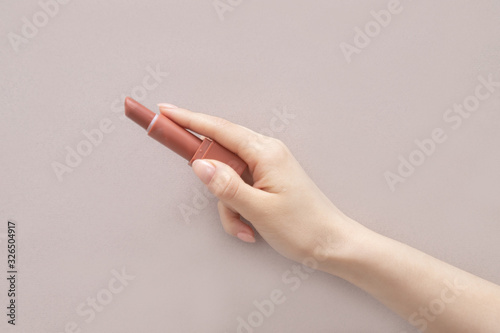 A girl's hand with a manicure holds a bright lipstick on a light background. Beige background. Red lipstick. Coral lipstick. Terracotta lipstick. Brick-colored lipstick. Makeup. Concept