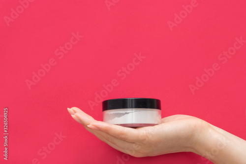 The hand of a girl with a manicure holds a jar of face powder on a pink background. White powder for the face. Transparent face powder. Makeup.