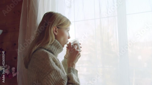 Woman standing in the window in a sweater and drinks hot cocoa. Girl enjoys a hot drink