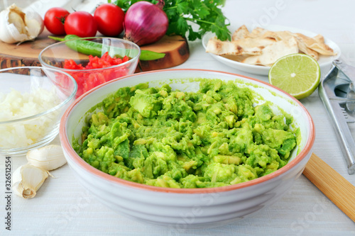 Mashed avocado along with diced onions and tomatoes  in bowls for making avocado dip. Preparing ingredients for guacamole with ingredients and kitchen utensils on table