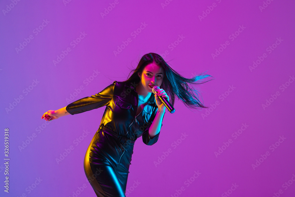 Caucasian female singer portrait isolated on purple studio background in neon light. Beautiful female model in black wear with microphone. Concept of human emotions, facial expression, ad, music, art. Caucasian female singer portrait isolated on purple studio background in neon light. Beautiful female model in black wear with microphone. Concept of human emotions, facial expression, ad, music, art.