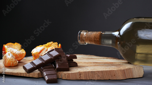 bottle of white wine with pieces of milk chocolate and tangerine on a wooden forest stand on a dark background. romantic evening dinner for two close-up