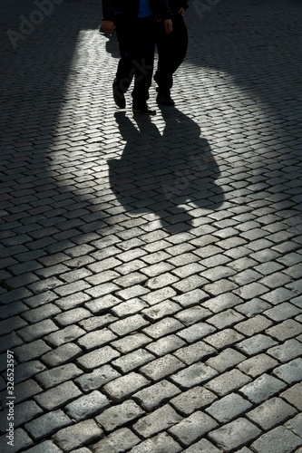 Silhouette view of two unrecognizable people walking together among shadows on a delicately lit cobblestone plaza