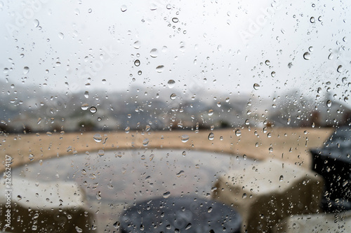 View from inside a hilltop home of a patio and subdivision below as rain falls. Taken through a rain covered and water spotted window .