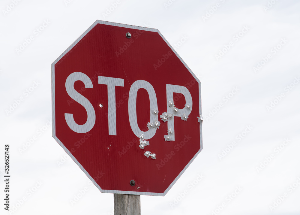 Red stop sign full of bullet holes with a background of white to grey clouds.  