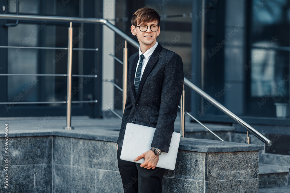 Fototapeta premium A handsome young businessman holds his laptop while standing in front of a glass building