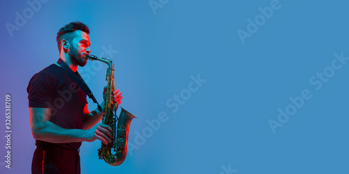 Young caucasian jazz musician playing the saxophone on gradient blue-purple studio background in neon light. Concept of music, hobby, festival. Joyful attractive guy. Colorful flyer for proposal.