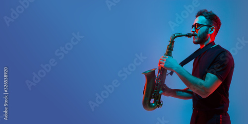 Young caucasian jazz musician playing the saxophone on gradient blue-purple studio background in neon light. Concept of music, hobby, festival. Joyful attractive guy. Colorful flyer for proposal.
