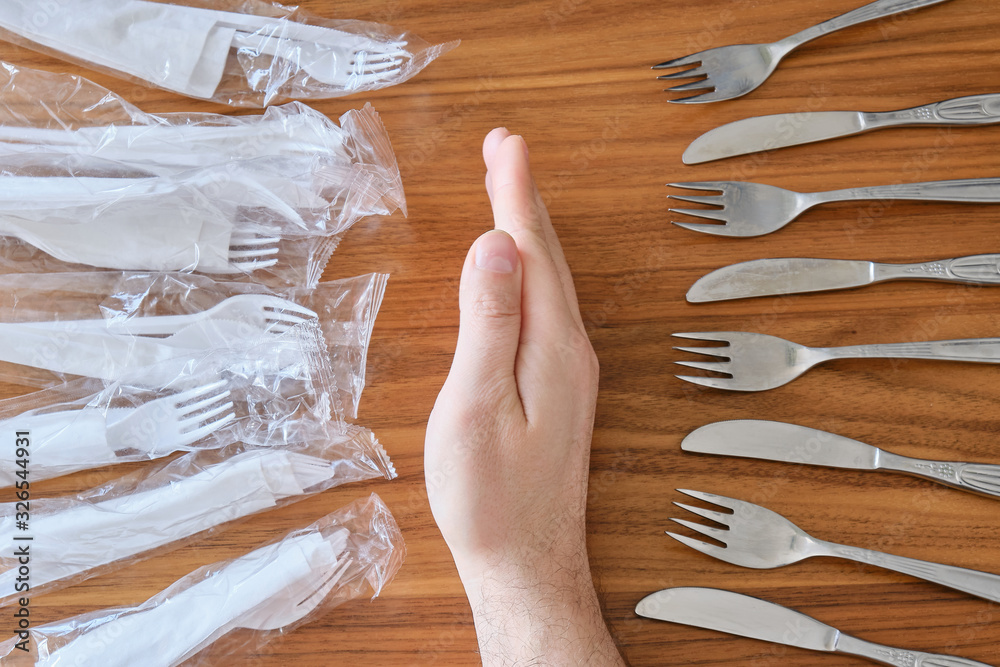 Plastic versus reusable cutlery concept: Man hand between disposable plastic sets and a stainless steel fork / knife set, on a walnut table top.