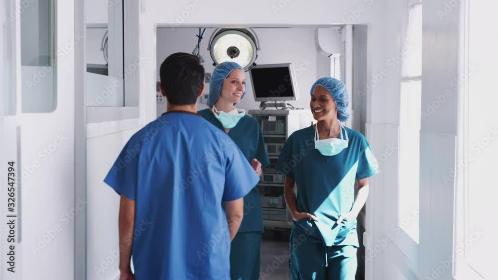 Two Female Surgeons Wearing Scrubs Having Meeting As They Walk Along Busy Hospital Corridor Together