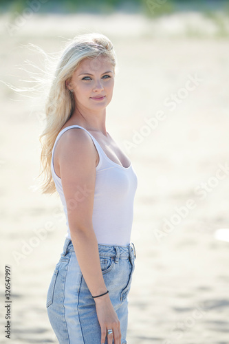 Stunning beautiful woman with blonde hair poses on beach in white tank top and denim skirt