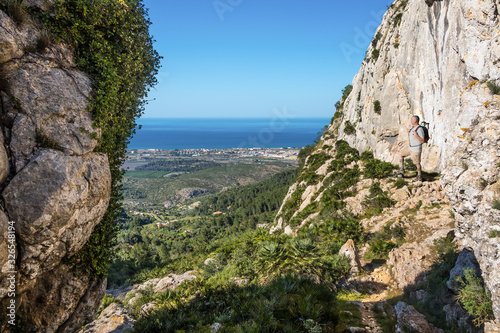 A view of the Mediterranean Sea from the mountain Segaria near the Spanish port city of Denia in sunshine and blue sky. On the right is a hiker with a backpack and looks into the valley.