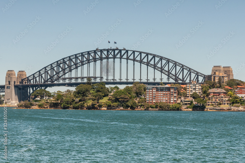 Fototapeta premium Sydney, Australia - December 11, 2009: Harbour Bridge, full metal span, bow and stone anchor towers against blue sky and above East shore with green vegetation and buildings. Azure bay in front.