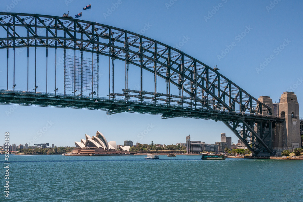 Naklejka premium Sydney, Australia - December 11, 2009: Opera House under Harbour Bridge, metal span, bow and stone anchor towers against blue sky and above azure bay water. Some boats.