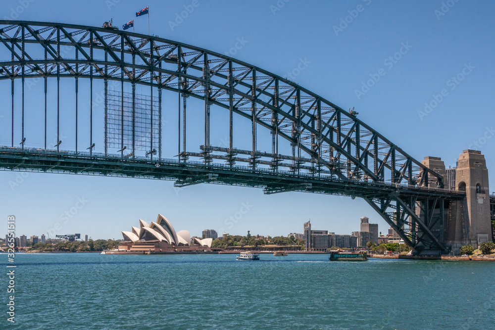 Sydney, Australia - December 11, 2009: Opera House under Harbour Bridge, metal span, bow and stone anchor towers against blue sky and above azure bay water. Some boats.