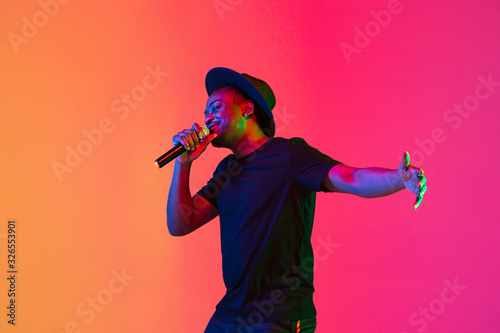 Young african-american musician singing on gradient orange-purple studio background in neon light. Concept of music, hobby, festival. Joyful party host, stand upper. Colorful portrait of artist.