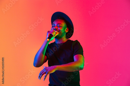 Young african-american musician singing on gradient orange-purple studio background in neon light. Concept of music, hobby, festival. Joyful party host, stand upper. Colorful portrait of artist. photo