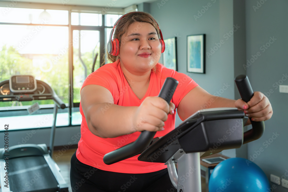 Happy Asian Overweight woman with earphone training on exercise bike in modern gym, happy and smile during workout. Fat woman take care of health and want to lose weight concept.. Happy Asian Overweight woman with earphone training on exercise bike in modern gym, happy and smile during workout. Fat woman take care of health and want to lose weight concept..