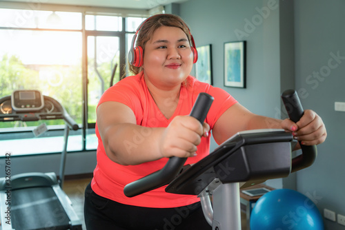 Happy Asian Overweight woman with earphone training on exercise bike in modern gym, happy and smile during workout. Fat woman take care of health and want to lose weight concept..