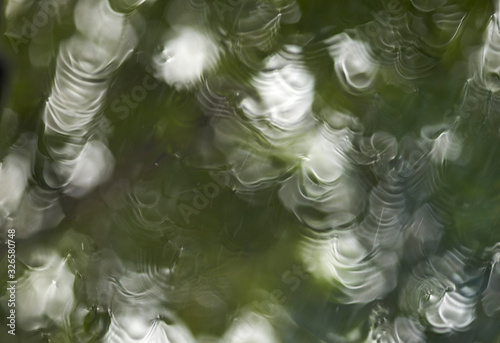 Looking through a window on a rainy day - rippled water, with streaks and droplets - along with dappled light from a green tree, combine in a blurry bokeh background image, themes of wet weather etc.