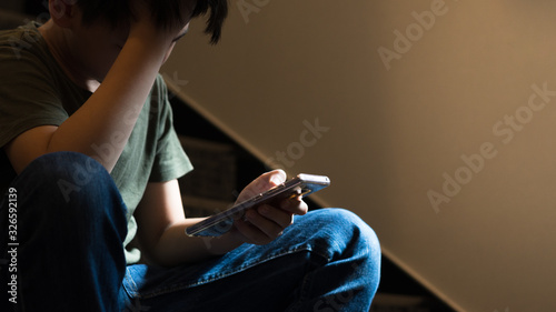Young Asian preteen/teenage boy sit on stair at home, cry and cover his face with hand, other hand hold smartphone, feeling alone, stressed, frustrated, overwhelmed, depressed. Cyber bullying concept. photo