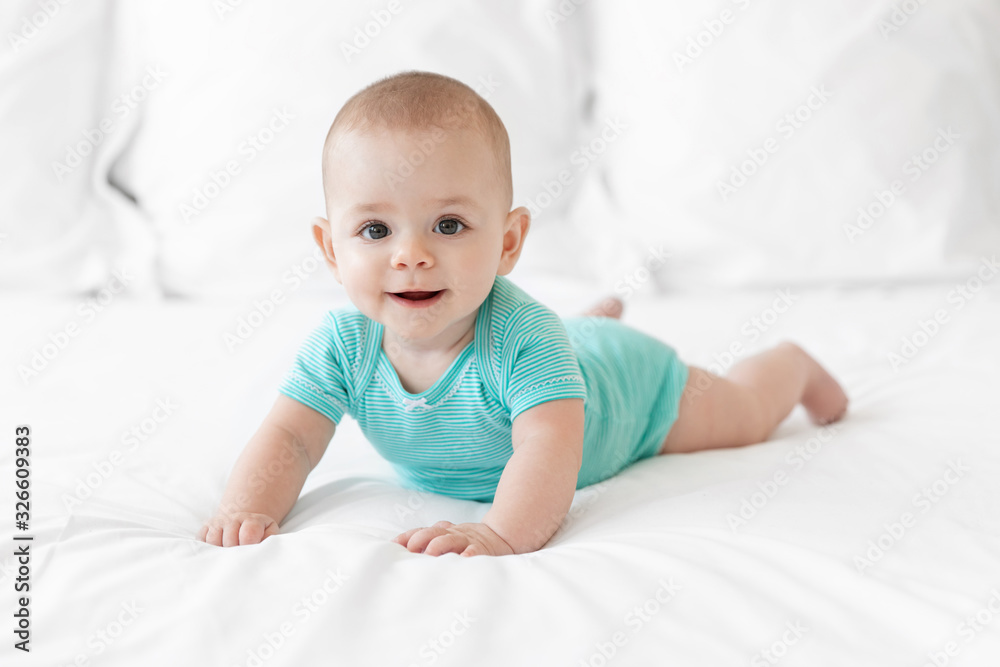 Happy baby in teal onesie lying on tummy on white bed Happy baby in teal onesie lying on tummy on white bed