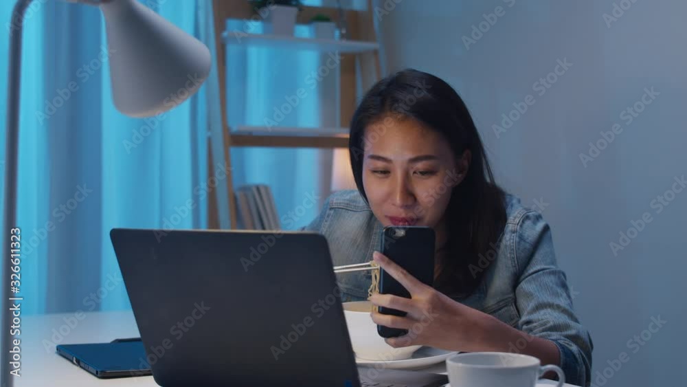 Freelance smart business women eating instant noodles live on social media via mobile phone in living room at home at night. Happy young Asian girl sitting on desk work overtime, enjoy relax time. Freelance smart business women eating instant noodles live on social media via mobile phone in living room at home at night. Happy young Asian girl sitting on desk work overtime, enjoy relax time.