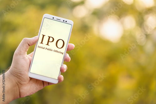 IPO Background Concept. Initial Public Offering concept. Young male hand holding mobile phone & showing IPO which is displayed on it's screen in outdoor background with copy space. photo