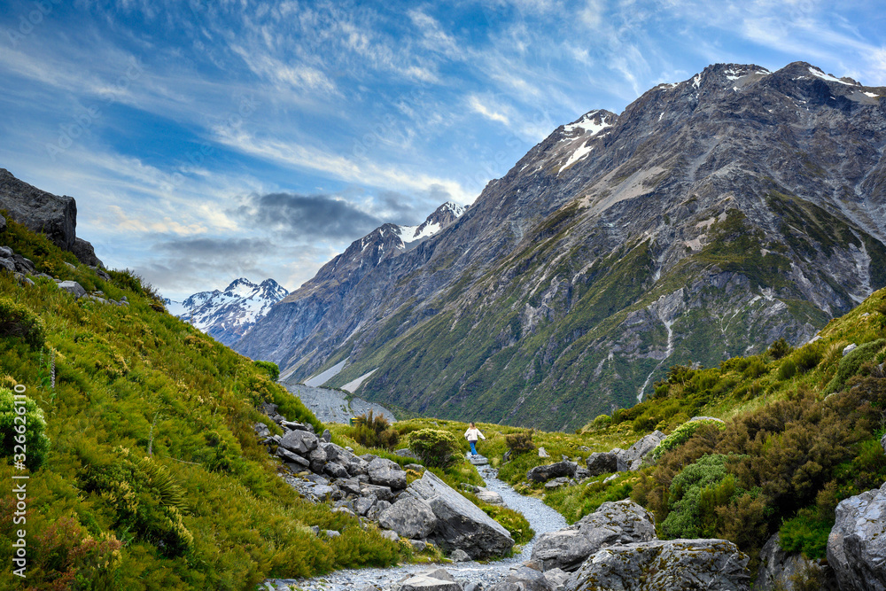 A female tourist walks the mountain pathway with a beautiful view of the high mountains above the snow and clouds. In the morning blue sky in Mount Cook National Park, Aoraki, New Zealand