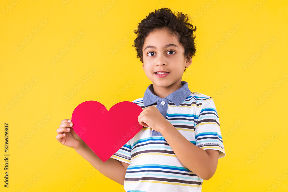 Curly pretty dark skinned boy in striped blue t-shirt is holding in hand red paper heart sign. Black cute child is smiling on yellow orange background. Emotional portrait concept. Saint Valentine day. Curly pretty dark skinned boy in striped blue t-shirt is holding in hand red paper heart sign. Black cute child is smiling on yellow orange background. Emotional portrait concept. Saint Valentine day.