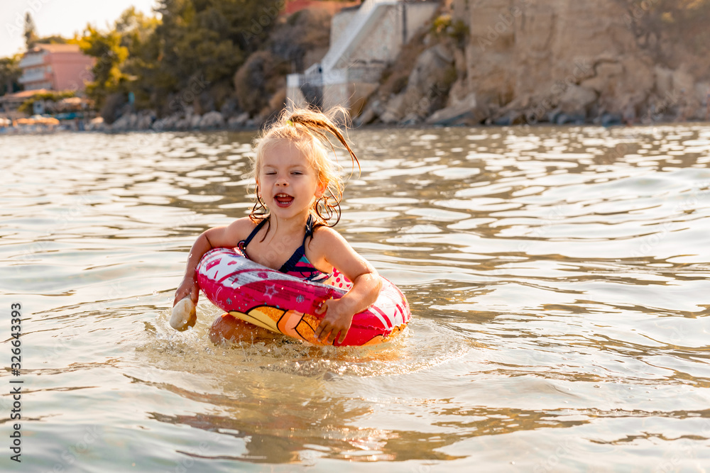 a little girl with a life buoy with a mischievous look jumps into the sea in the rays of the setting sun, in the hands of a large stone, a flare on her hair. summer holiday concept a little girl with a life buoy with a mischievous look jumps into the sea in the rays of the setting sun, in the hands of a large stone, a flare on her hair. summer holiday concept