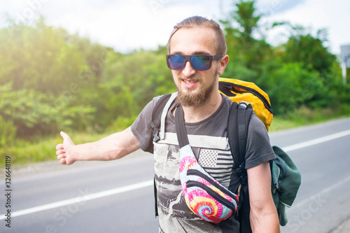 Young man in trendy standing in the middle of highway and looking away. Full-length portrait of traveler with big backpack walking down the road in morning. Emotional traveler in adventure.