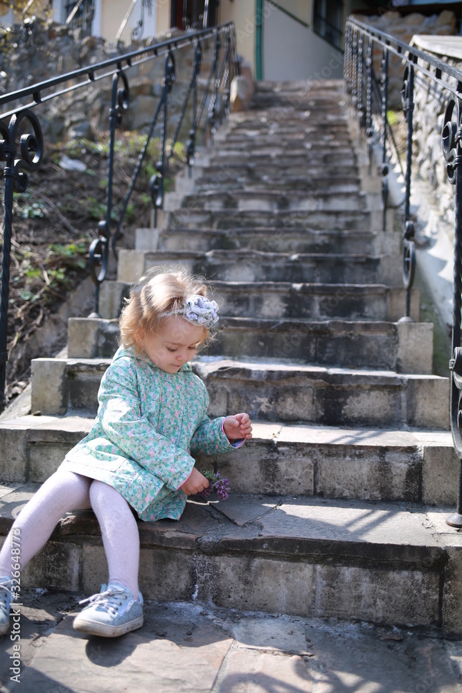 A little girl sits on the steps in the spring botanical garden with an apple in her hands and eats it where the primroses bloomed A little girl sits on the steps in the spring botanical garden with an apple in her hands and eats it where the primroses bloomed