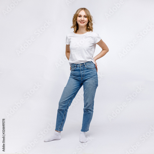 A blonde girl is stands and smiling in a white t-shirt and blue jeans on a white background.