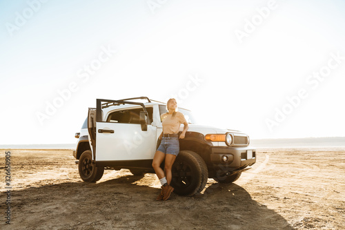 Beautiful young girl standing at the car at the sunny beach photo