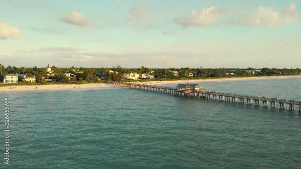 Naples Beach and Fishing Pier at Sunset, Florida. Drone flies back. Drone flies back, flying away from pier. Drone aerial of Naples coastline into Naples fishing pier with the sunset in the background Naples Beach and Fishing Pier at Sunset, Florida. Drone flies back. Drone flies back, flying away from pier. Drone aerial of Naples coastline into Naples fishing pier with the sunset in the background