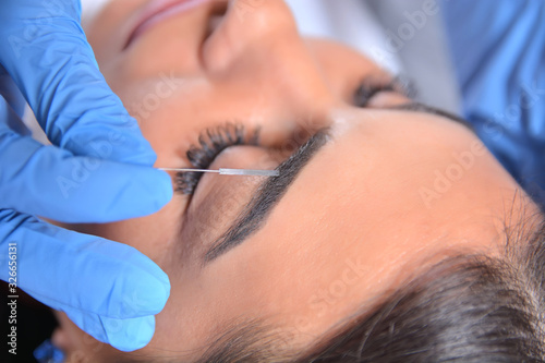 Young pretty girl having treatment on eyebrows in beautician salon. Cosmetologist tattooing with the professional and sterile little metal tool. Close up, selective focus