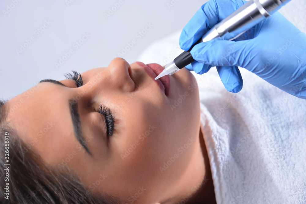 The procedure of professional applying permanent makeup on woman lips. Young beautiful girl getting a tattoo in a beauty salon. Beautician hand with a professional tool. Close up, selective focus The procedure of professional applying permanent makeup on woman lips. Young beautiful girl getting a tattoo in a beauty salon. Beautician hand with a professional tool. Close up, selective focus
