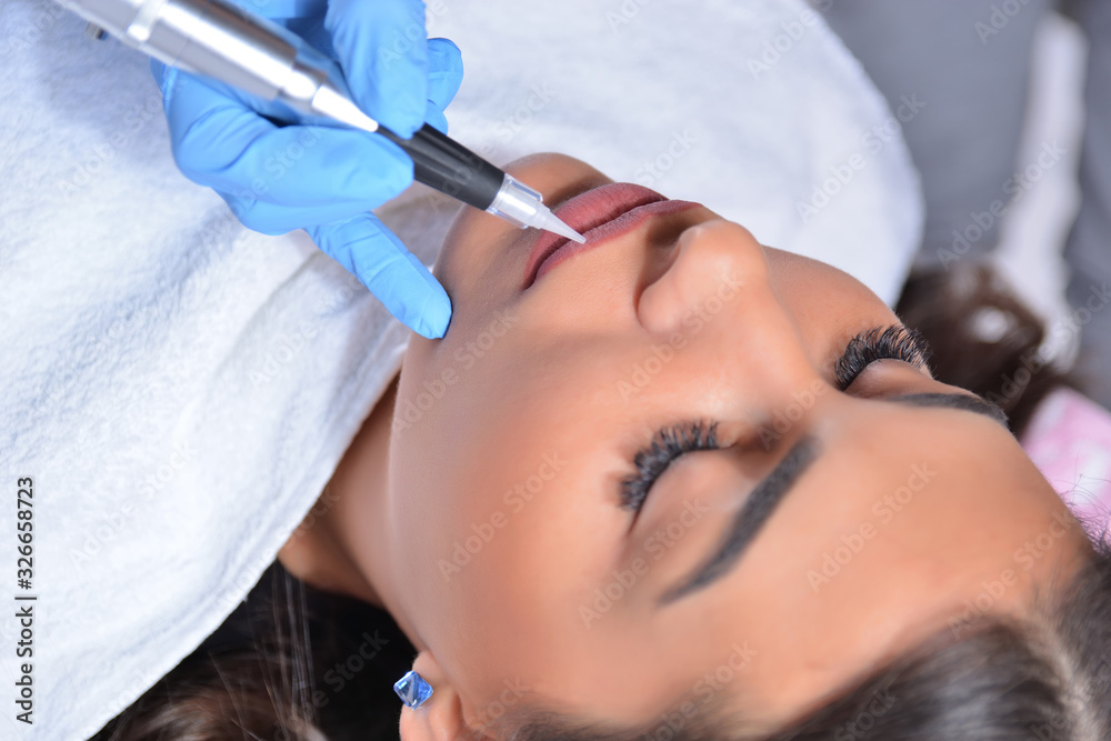 Naklejka premium Young pretty girl having permanent makeup on lips in beautician salon. Cosmetologist applying tattoo with the professional and sterile tool. Close up, selective focus