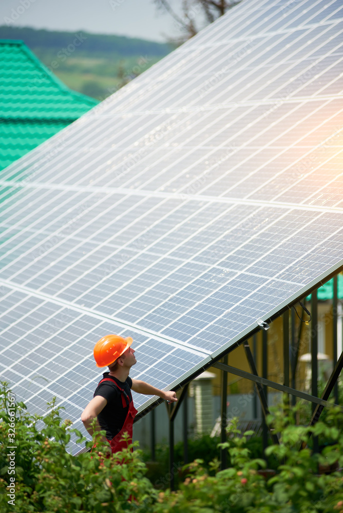 Young man in an orange helmet near a high solar station on a plot of land. Alternative cheap sun energy production and profitable financial investment concept
