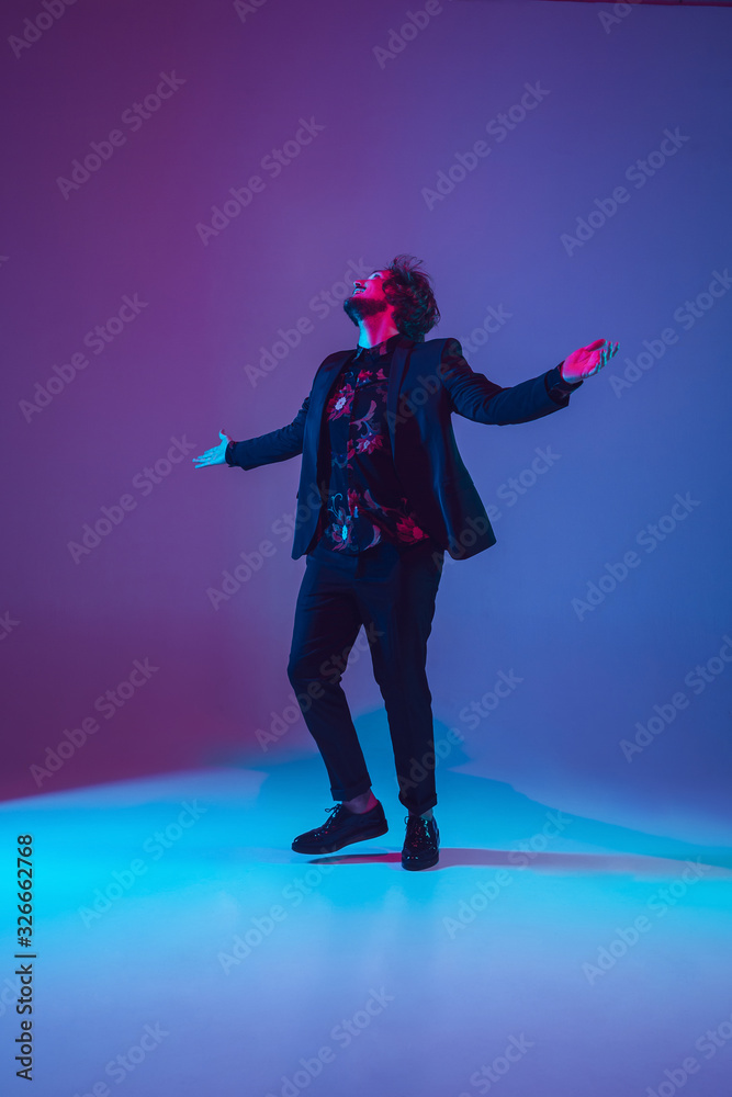 Obraz premium Young caucasian musician in casual dancing on gradient blue-purple background in neon light. Concept of music, hobby, festival. Joyful party host, DJ, stand upper, dancer. Colorful portrait of artist.