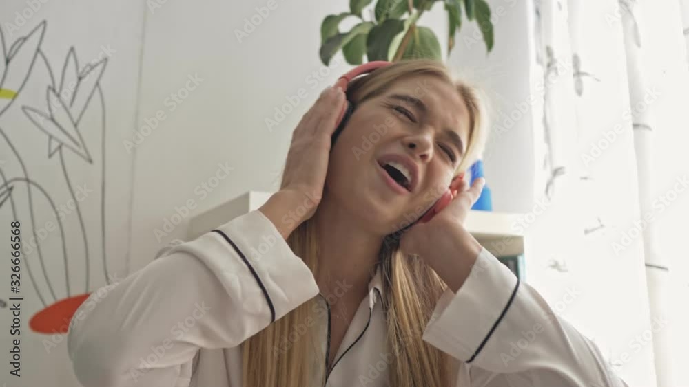 A pretty young woman wearing white pajamas is listening to music and dancing in the bedroom A pretty young woman wearing white pajamas is listening to music and dancing in the bedroom