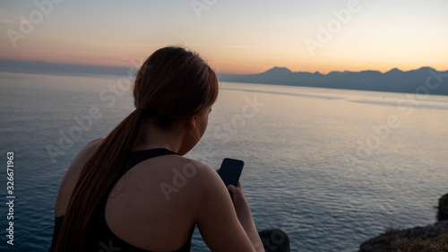 Young sports girl using mobile phone after exercise by the sea over huge cliffs at sunset. She wears tights and sports bra.