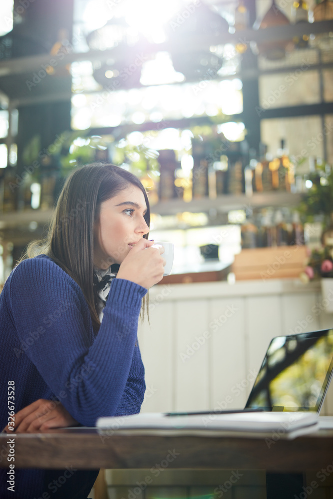 Attractive caucasian in sweater sitting in cafe, holding coffee and looking away. On table is laptop. Attractive caucasian in sweater sitting in cafe, holding coffee and looking away. On table is laptop.