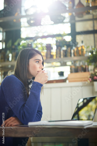 Attractive caucasian in sweater sitting in cafe, holding coffee and looking away. On table is laptop.