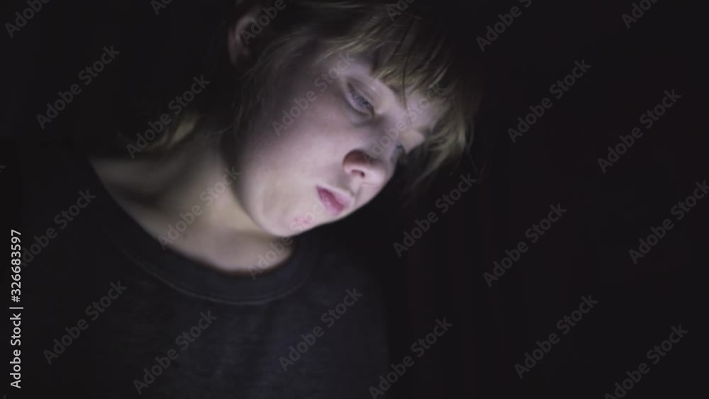 Sad/depressed teenager sitting on her bed in the dark while scrolling through her phone. The light from the phone is illuminating her face. Sad/depressed teenager sitting on her bed in the dark while scrolling through her phone. The light from the phone is illuminating her face.