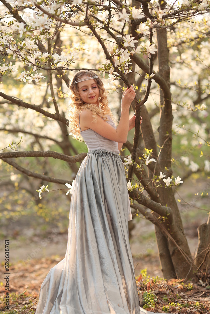 young beautiful blonde in a silver dress with a train in the spring garden, where white magnolia blooms